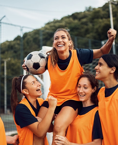 Estudantes sorridentes levantando sua colega após uma partida de futebol.