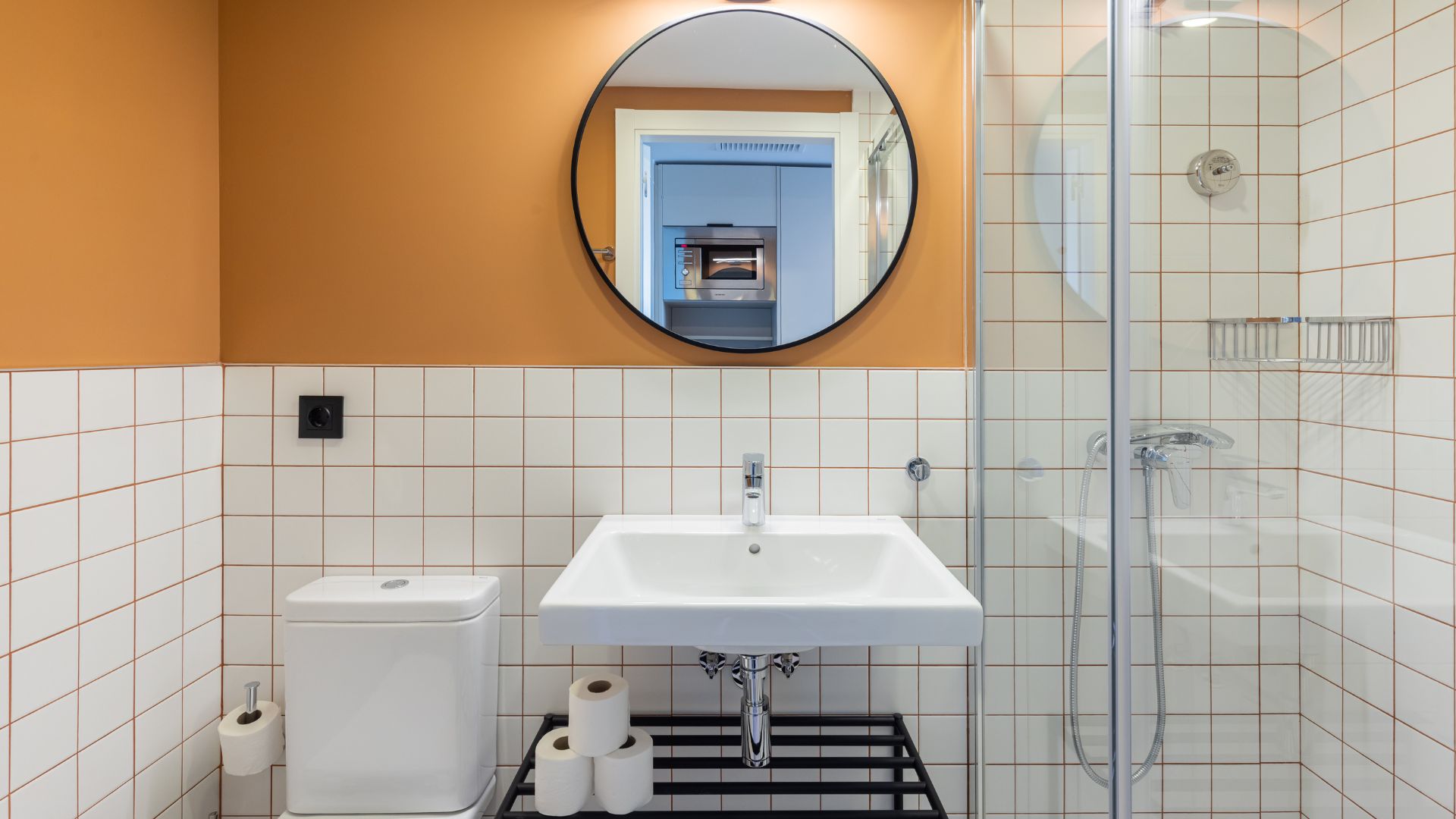 Baño completo del estudio con ducha, lavabo, espejo redondo y azulejos en tonos neutros y cálidos.