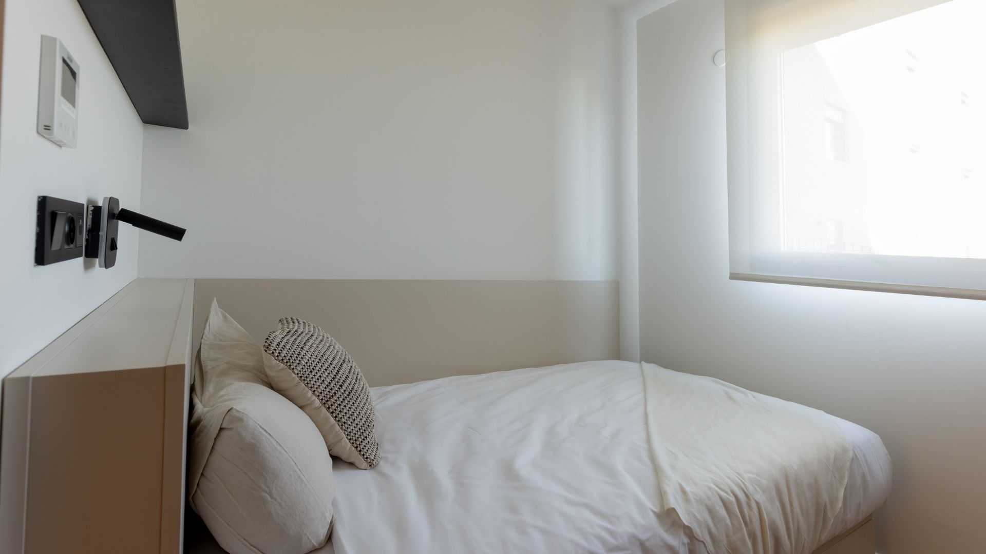 Dormitorio de estudio con cama individual, estantería superior, iluminación de lectura y diseño funcional.